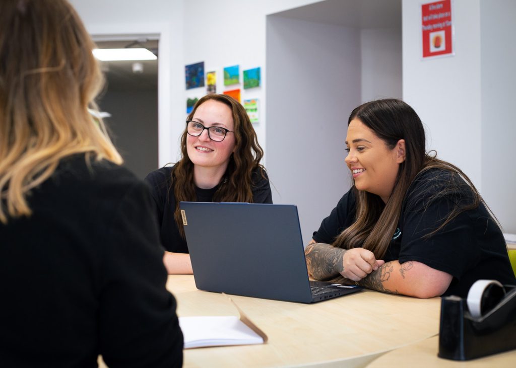 An image of 2 women in the beyond the margin team sat at a desk having a conversation whilst looking at laptop