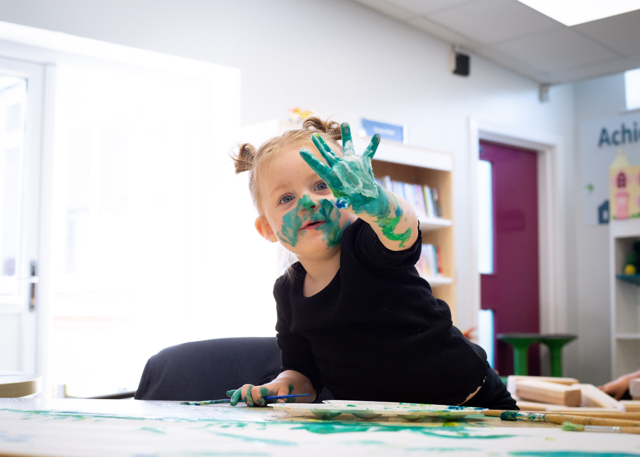 A smiling girl toddler holds up her painted hand to the camera